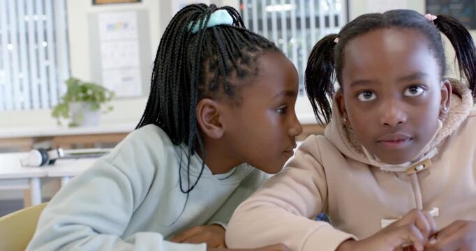 African teen female students leaning in sharing secret at class desk mint top tan hoodie glancing