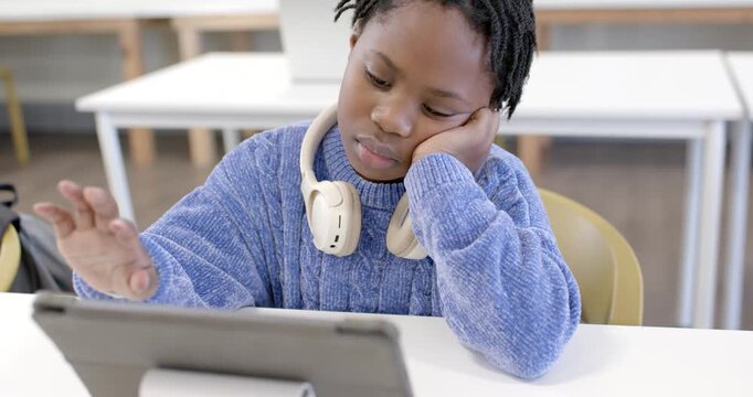 African youth female sitting in study area tapping tablet after prompt, wearing headphones to study