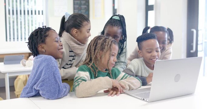 Diverse schoolchildren leaning around silver laptop at classroom table, reacting to laptop content