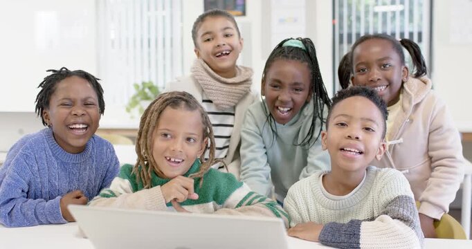 Diverse children watching laptop for class activity leaning forward and laughing at white table
