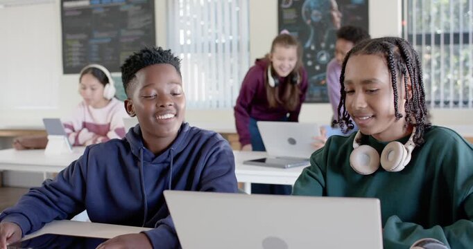 Two teenage boys exchanging glances, working on laptop in classroom, right teen pointing and typing
