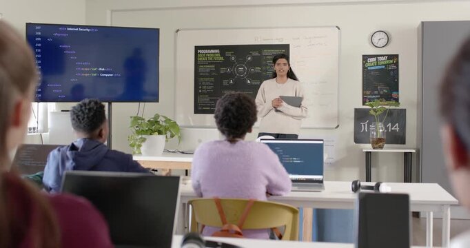 Indian woman teacher starting class holding tablet pointing at code guiding students on laptops