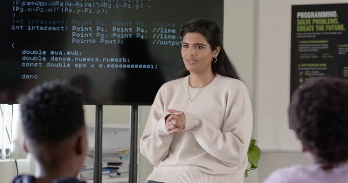 Indian female mentor on stool pointing to code on screen, guiding African American youth in coding