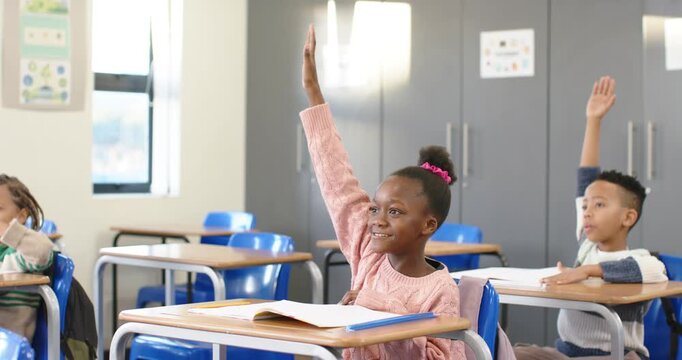 African American child in pink sweater raising hand answering teacher prompt at desk with workbook