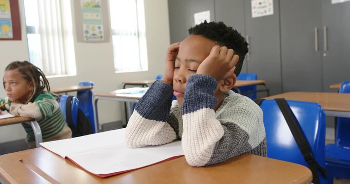 African American classmates watching boy at desk in class losing focus and laying head on notebook