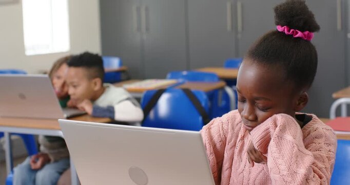 African American kid in pink sweater frowning, leaning, typing on silver laptop at desk, copy space