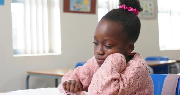 Leaning over desk African child pink sweater holding yellow pencil writing pausing doing classwork