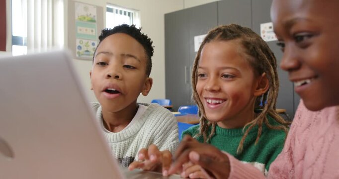 Diverse children leaning over laptop at school desk, reacting to on-screen task, typing classwork