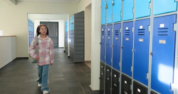 Youth African American female walking school hallway glancing at blue locker storing green backpack
