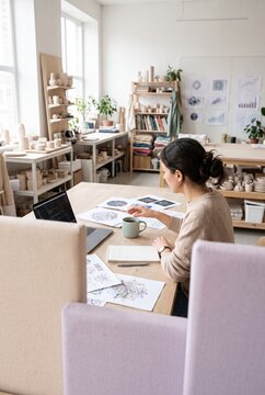 Young female artist and designer analyzing data charts and working on a laptop at a wooden desk in a pottery studio workspace