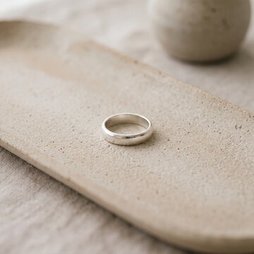 Minimalist silver wedding ring resting on a textured ceramic tray over a neutral linen surface, showcasing modern minimalist jewelry design