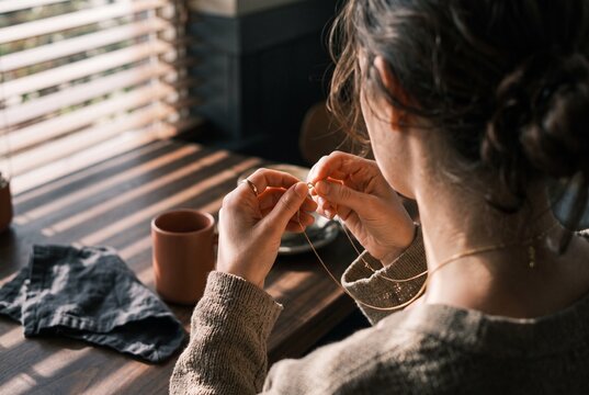 Young woman wearing a delicate gold necklace while sitting at a wooden table in warm morning sunlight near a window with blinds