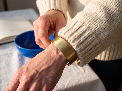 A close-up shot of a woman wearing a broad gold brass cuff bracelet on her wrist over a cozy white knit sweater