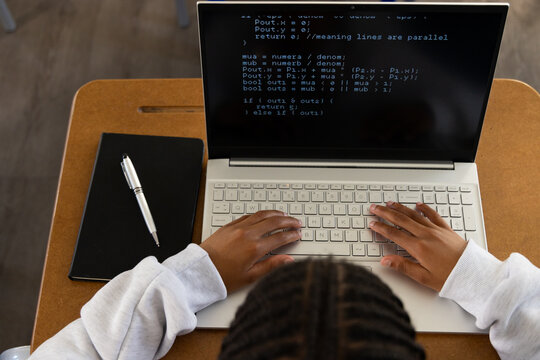 African American teen male typing code on silver laptop at wooden student desk with notebook pen