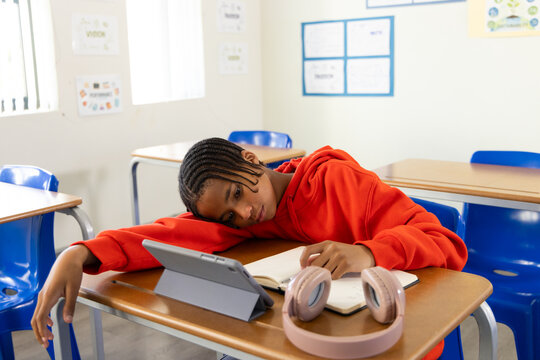 African American teen student in red hoodie leaning on forearm at school desk with tablet, headset