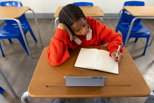 African male teenage student in red hoodie taking notes at school desk with tablet pink headset