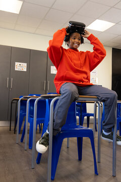 African American male youth sitting on desk in classroom wearing red hoodie holding VR headset