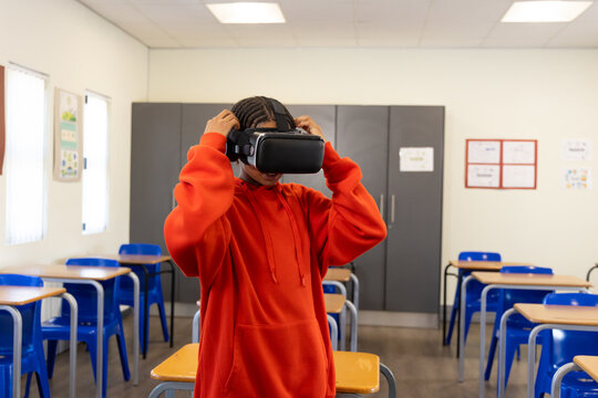 African youth male student standing between desks in classroom, adjusting VR headset in red hoodie