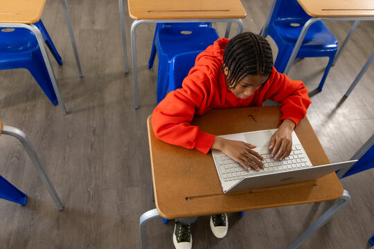 African American teen student typing on silver laptop at school desk with pencil groove, red hoodie