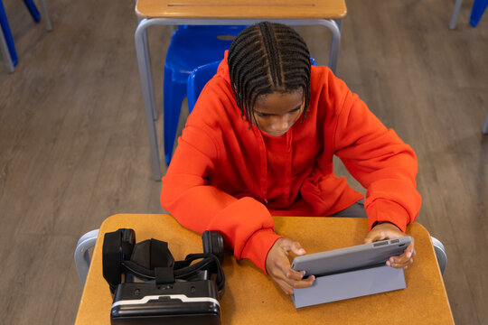 African American youth wearing red hoodie sitting at school desk using tablet, VR headset resting