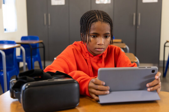 African American youth in orange hoodie sitting at school desk holding tablet VR headset resting