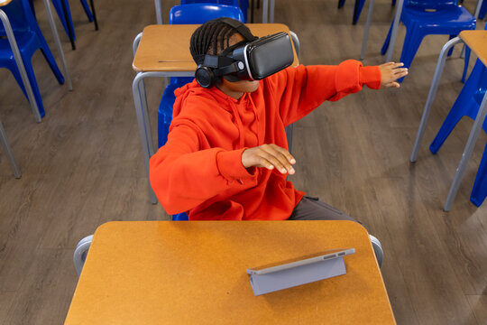 African American teen in orange hoodie using VR headset and headphones at school desk with tablet