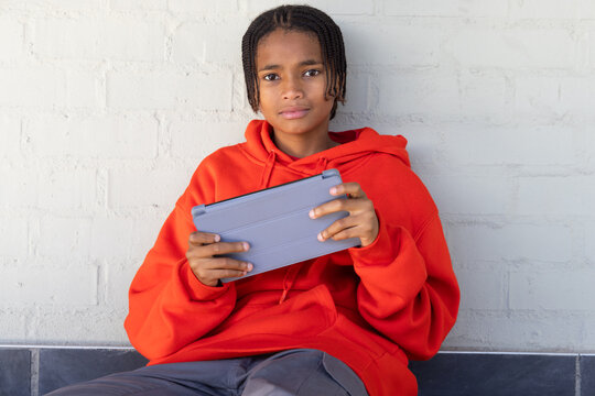 Teen male sitting against gray brick wall holding gray tablet with folio-cover wearing red hoodie