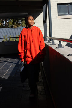 Youth male wearing bright red oversized hoodie standing on covered walkway holding briefcase