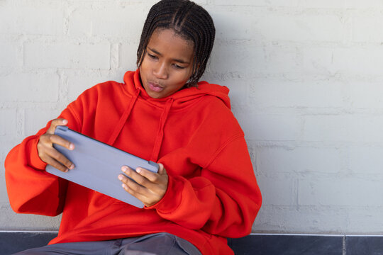 African American teenager in red hoodie sitting against gray wall on dark ledge holding gray tablet
