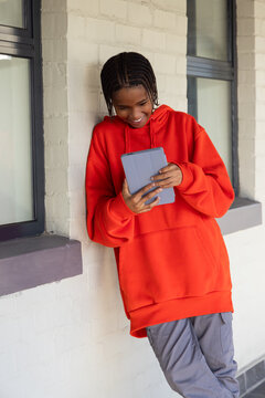 Youth leaning against white brick wall outside wearing red hoodie holding tablet with folio