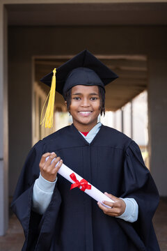 African American youth male posing in portico with columns, holding diploma, wearing gown and cap