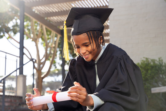 African youth male sitting steps under pergola in gown and cap with yellow tassel holding diploma