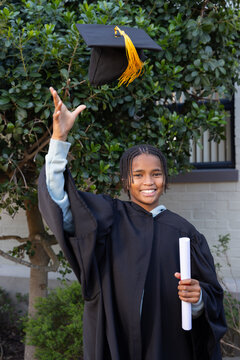 African American teenage male in black gown by brick wall tossing yellow tassel cap with diploma