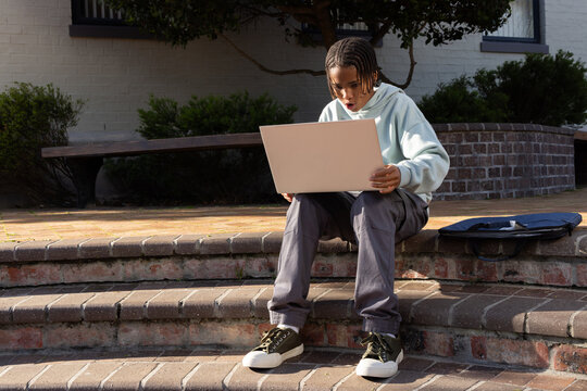 Teenage African American male sitting on front brick steps holding open laptop, backpack beside