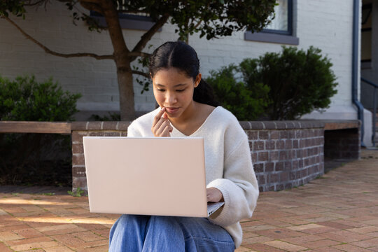 Asian female child sitting on brick paving at front porch holding laptop near low brick planter