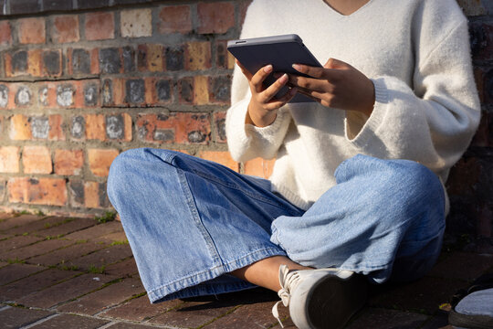 Child girl wearing cream knit sweater wide leg jeans holding and tapping dark tablet on bricks