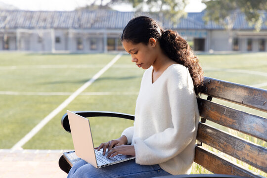 Female child wearing white sweater, jeans sitting on bench at sports field typing on silver laptop