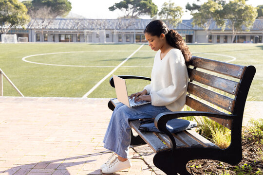 Child girl sitting on wooden bench at edge of soccer pitch typing on laptop, copy space