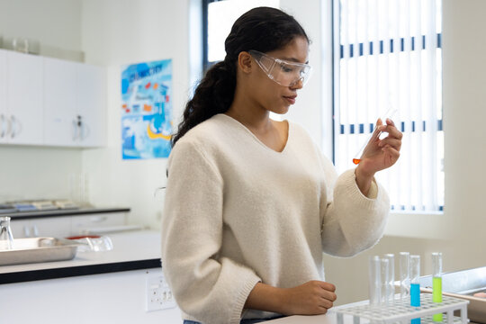 African-American teen wearing goggles holding pipette checking orange solution near tubes at school