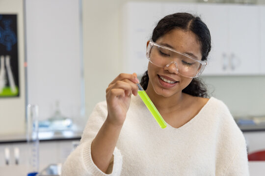 Child girl wearing goggles and sweater holding and inspecting neon green test tube at school lab