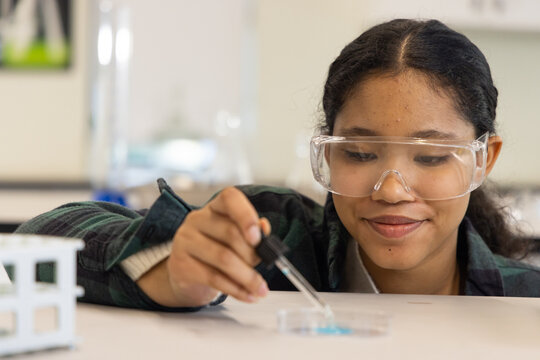 Female child in flannel and goggles using dropper above petri dish with blue-liquid at science lab