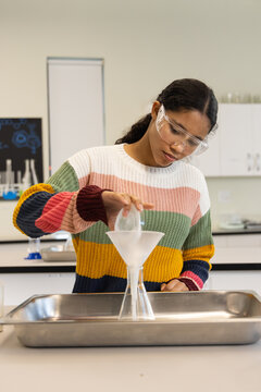 Child wearing goggles, striped sweater, pouring from beaker through funnel into flask at lab bench