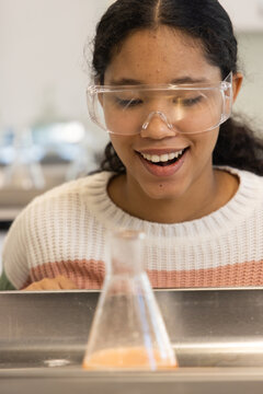 Girl wearing safety goggles and white sweater leaning at school lab sink observing orange flask
