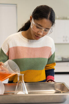 Child wearing goggles, striped sweater pouring orange liquid from pitcher into flask in lab
