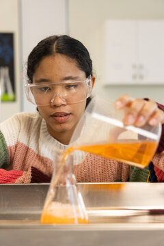 Female child wearing knit sweater, goggles pouring orange liquid from beaker into flask at lab sink