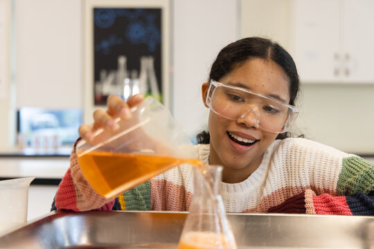 Child wearing goggles, bright sweater pouring orange solution from beaker into conical flask in lab