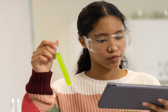 Child wearing colorful sweater and goggles holding neon green test tube and tablet in school lab