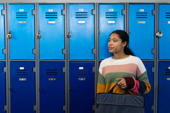 Asian child standing at school lockers locked, holding gray bag wearing striped sweater, copy space