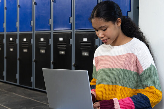Female child in sweater, typing laptop while leaning on right wall, school lockers, copy space