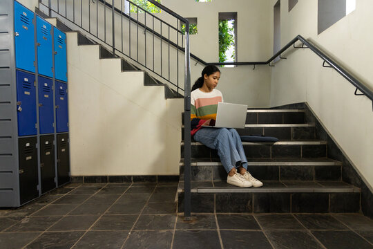 Female child sitting on tiled stair landing in school corridor using laptop near blue lockers